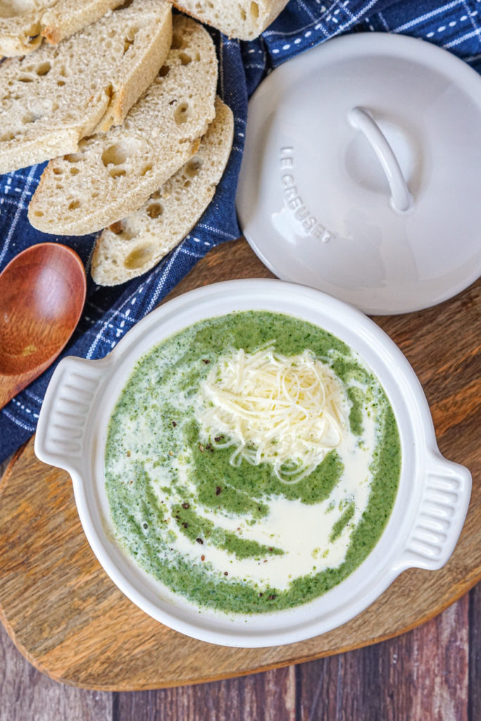 Spinach Gruyere Soup Aerial view of Spinach Gruyere Soup in a white soup bowl with cover on a wooden board next to slices of bread and a wooden spoon.