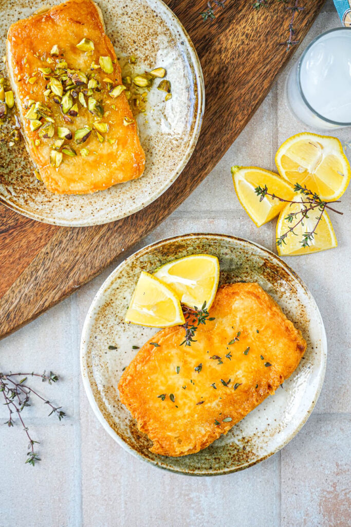 Aerial view of Saganaki (Greek Fried Cheese) on two plates, one with lemon wedges and thyme leaves and the other with pistachios and honey.