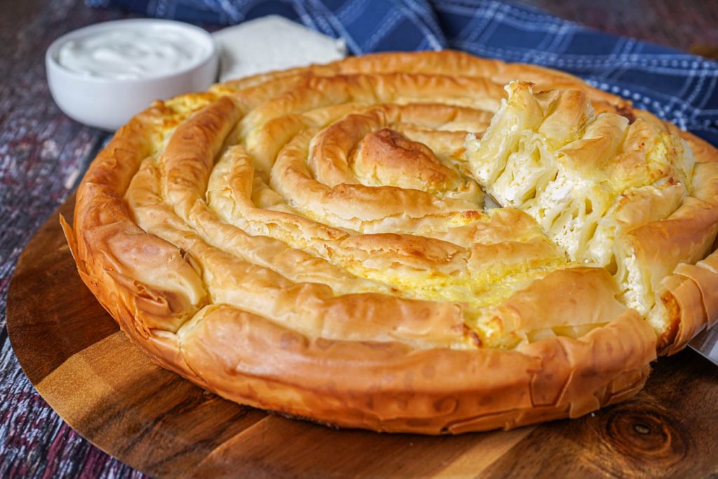 Banitsa (Bulgarian Cheese Pie) on a wooden board with a slice lifted up.