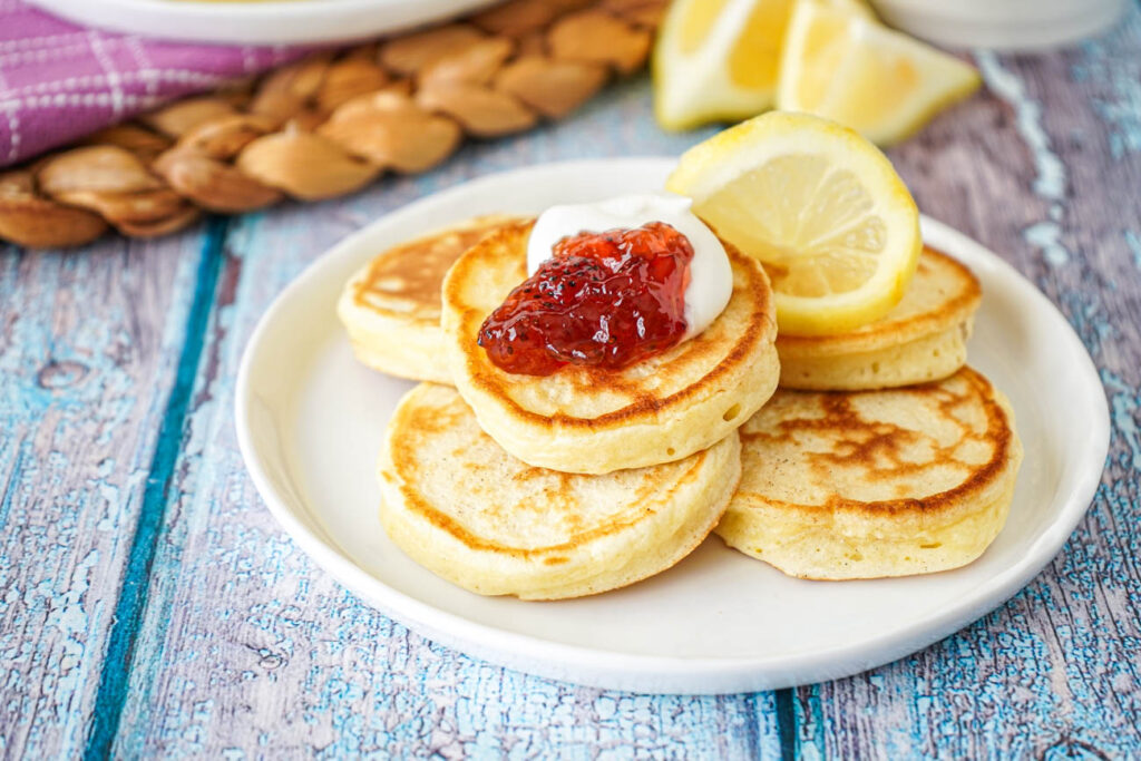 Five Lemon Pikelets on a white plate with strawberry jam, whipped cream, and a lemon slice.