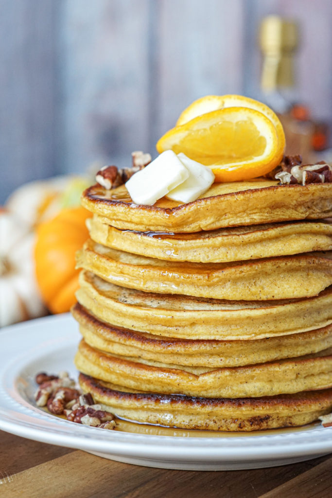 A stack of seven Orange Pumpkin Pancakes on a white plate and topped with orange slices.