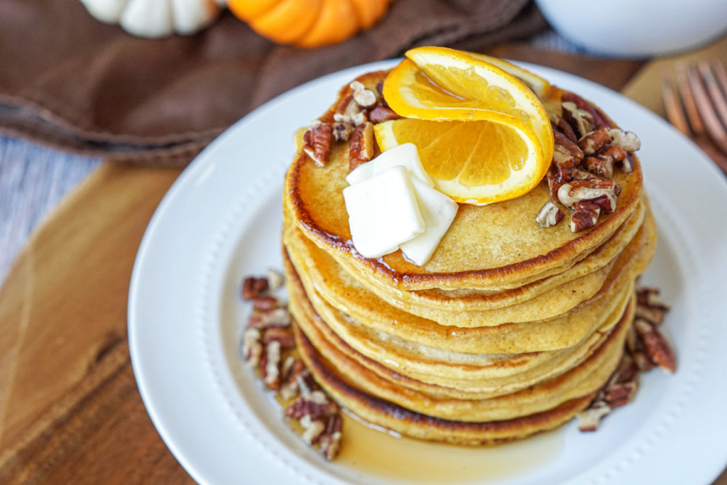 A stack of Orange Pumpkin Pancakes on a white plate and topped with orange slices and chopped pecans.