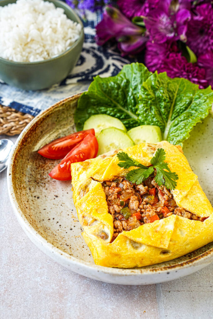 Close up of Kai Yad Sai (Thai Stuffed Omelette) on a plate with rice and purple flowers in the background.