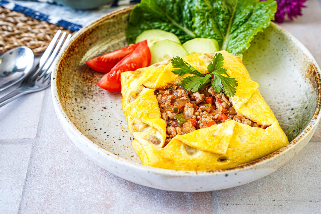 Kai Yad Sai (Thai Stuffed Omelette) in a bowl with lettuce leaves, cucumber slices, and tomato wedges.