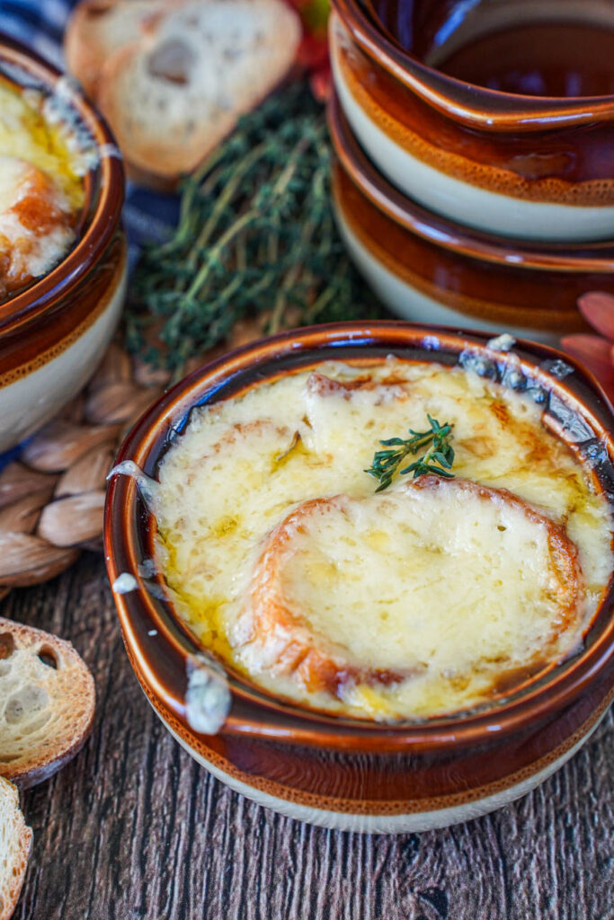 Close up of Vegetarian French Onion Soup in a brown bowl with more brown bowls and thyme leaves in the background.