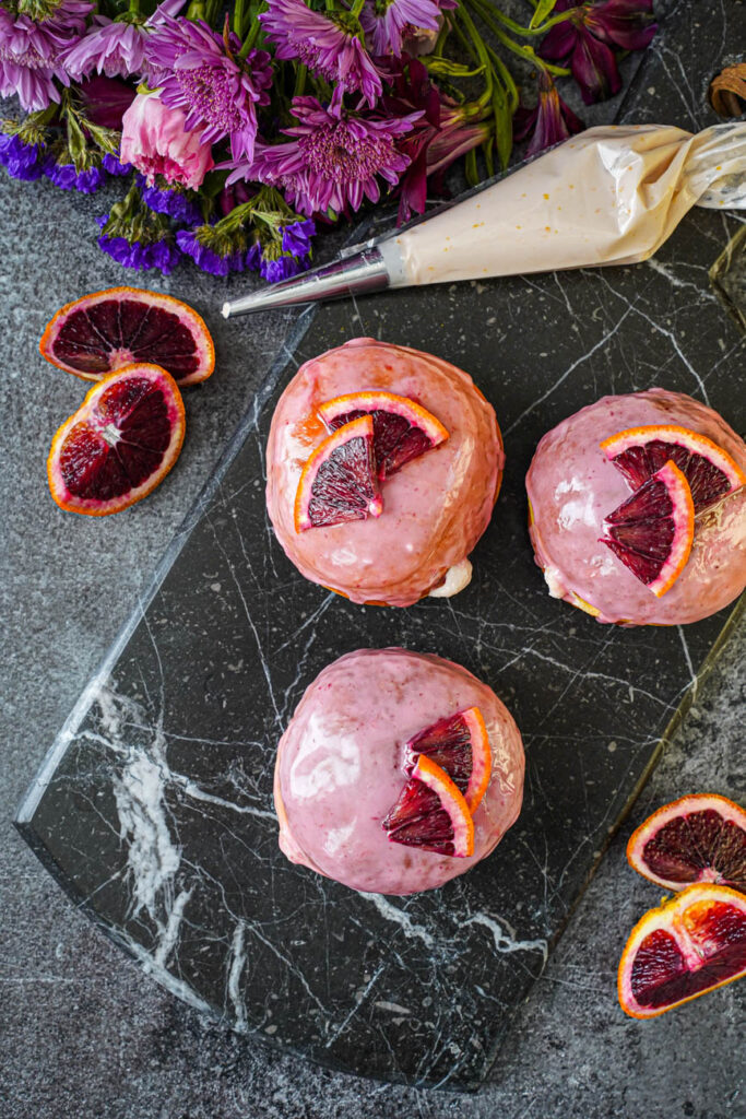 Aerial view of three Blood Orange Cream Cheese Doughnuts on a marble board next to a piping bag of blood orange cream cheese.