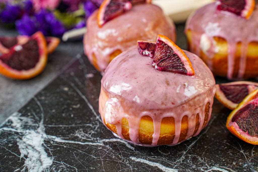 Three Blood Orange Cream Cheese Doughnuts on a dark marble board next to blood orange wedges.