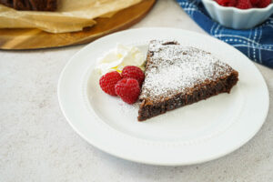 A slice of Kladdkaka (Swedish Chocolate Sticky Cake) on a plate with cream and raspberries.