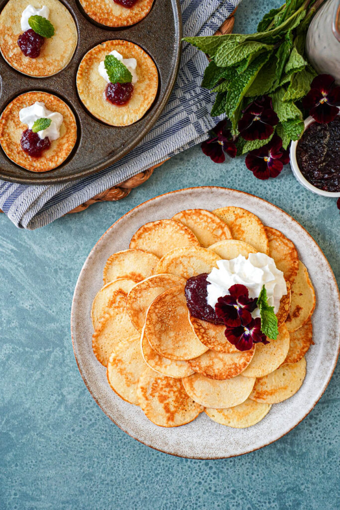 Aerial view of Plättar (Swedish Mini Pancakes) on a plate next to more pancakes in a pan, a bunch of mint, and red pansies.