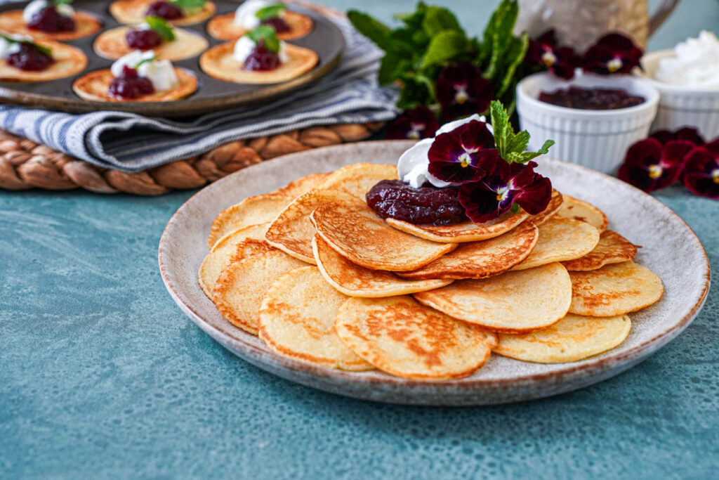 Plättar (Swedish Mini Pancakes) piled on a plate with pansies, lingonberry jam, and whipped cream.