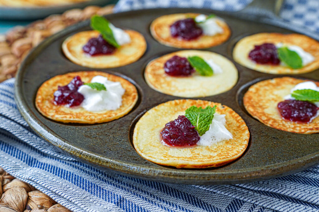 Plättar (Swedish Mini Pancakes) in a pan with dollops of lingonberry jam and whipped cream and a mint leaf on top.