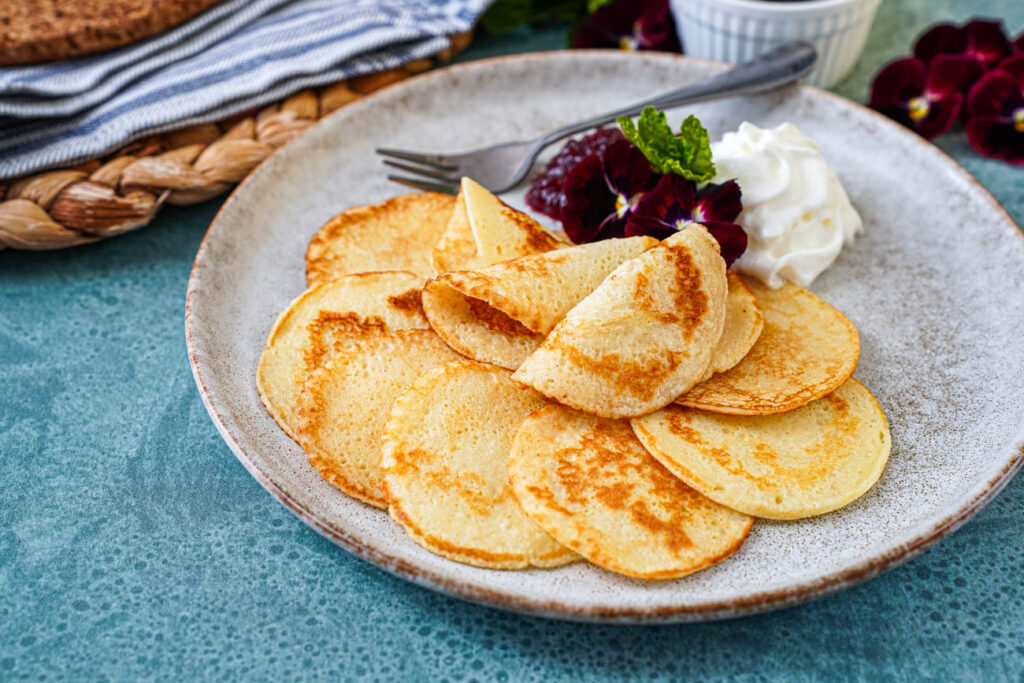 Plättar (Swedish Mini Pancakes) on a plate with pansies, whipped cream, lingonberry jam, and a fork.