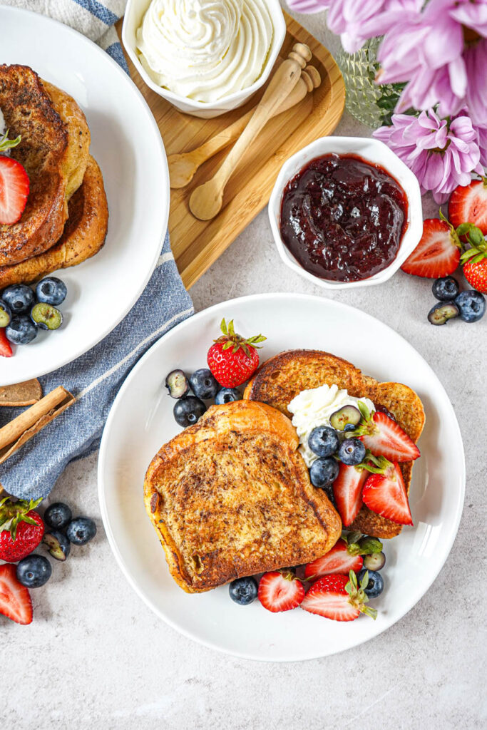 Aerial view of Arme Riddere (Norwegian Cinnamon Toast) on a two plates next to bowls of jam and whipped cream.