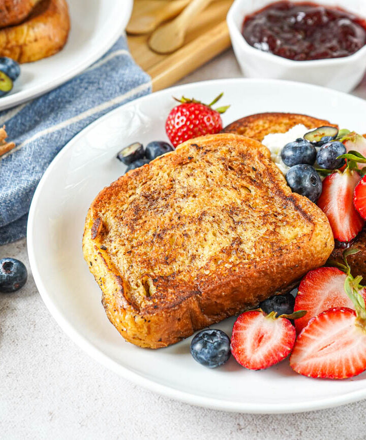 Two slices of Arme Riddere (Norwegian Cinnamon Toast) on a white plate with strawberries and blueberries.