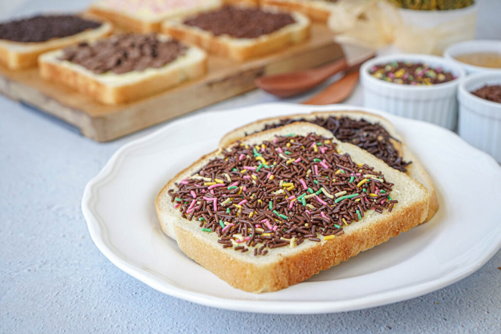 Two slices of Broodje Hagelslag (Dutch Bread with Sprinkles) on a white plate with more on a wooden board.