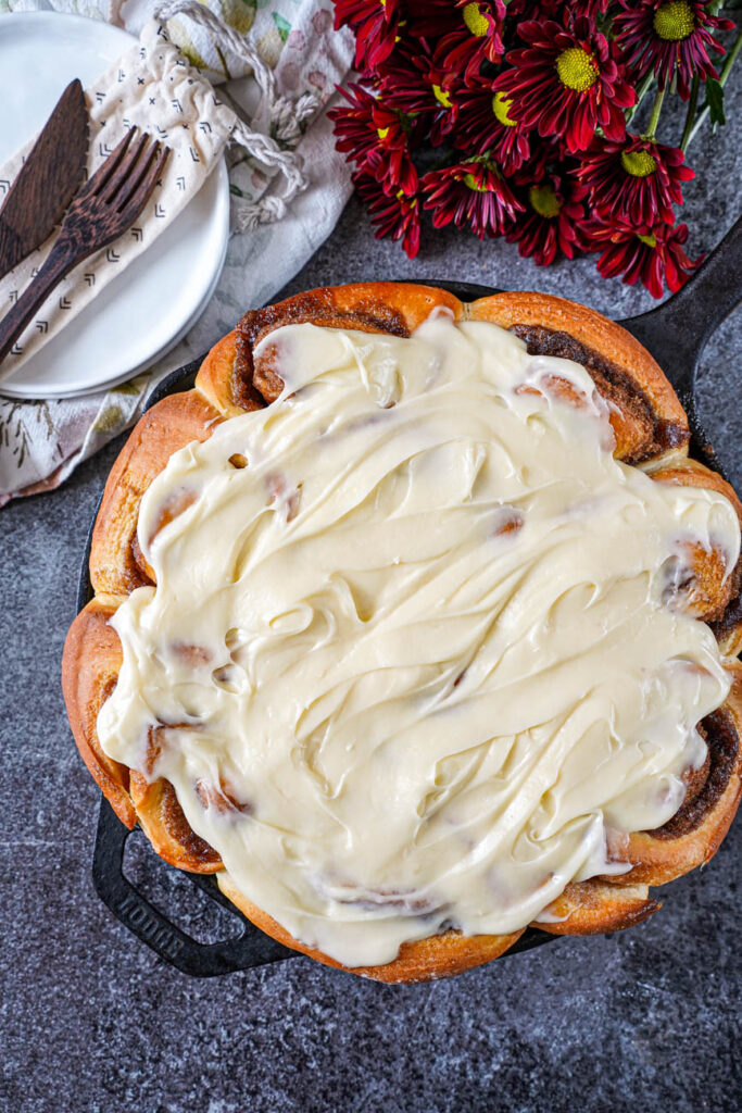 Aerial view of Cast Iron Cinnamon Rolls next to plates, wooden fork and spoon, and maroon flowers.