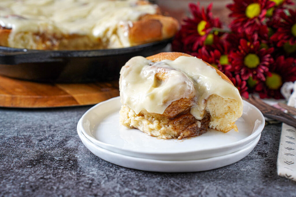 Cinnamon roll on a plate with more in a cast iron pan in the background.