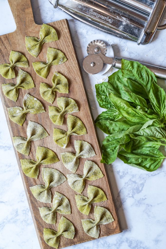 Aerial view of Homemade Basil Farfalle on a wooden board next to basil leaves and a pasta machine.