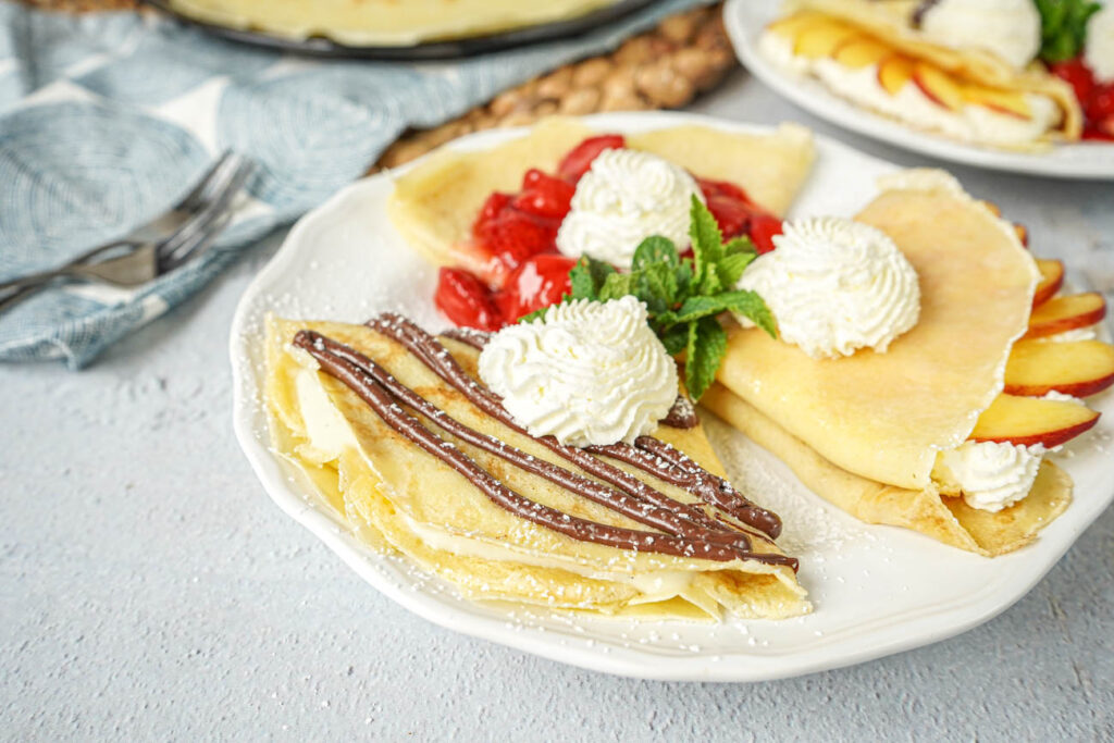 Sweet Crepe Trio on a white plate with peaches, Nutella, strawberries, whipped cream, and mint.