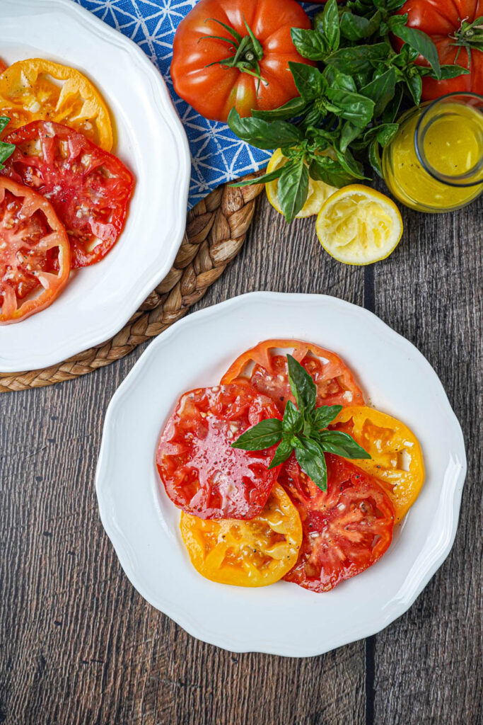 Aerial view of Tomato Salad with Lemon Dijon Dressing on two white plates next to tomatoes, basil, and lemon halves.
