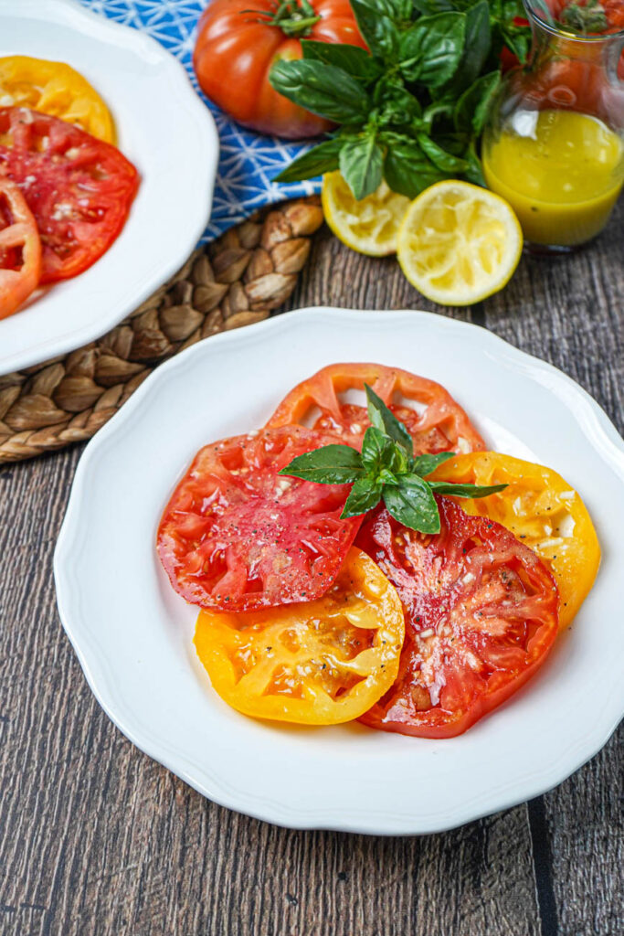 Tomato Salad with Lemon Dijon Dressing on two white plates with dressing in a glass, lemon halves, basil, and tomatoes in the background.