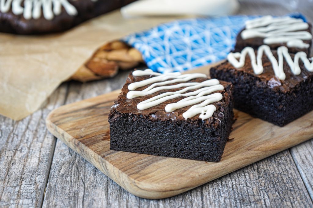 Three Dark Chocolate Brownies with a white chocolate drizzle on a wooden board.