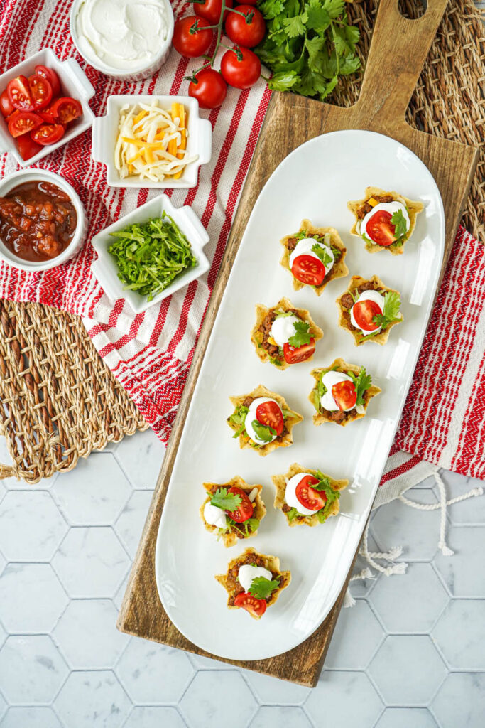 Aerial view of Mini Taco Bites on a white platter next to tomatoes, cilantro, cheese, lettuce, and sour cream.
