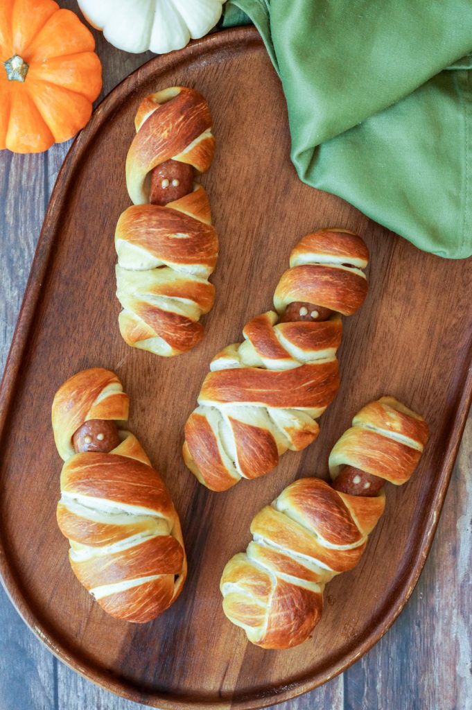 Aerial view of Mummy Pretzel Dogs on a wooden board next to pumpkins and green towel.
