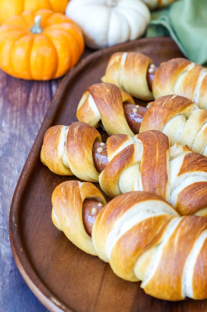 Close up of four Mummy Pretzel Dogs on a wooden board next to a green towel and orange and white pumpkins. 
