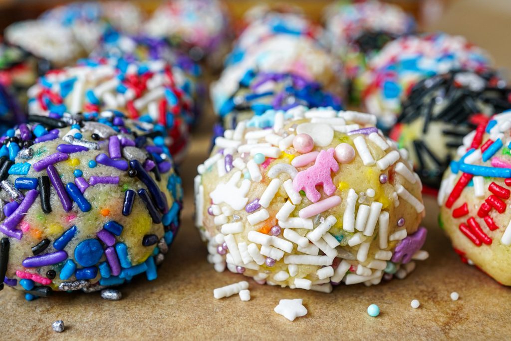 Small balls of cookie dough coated in sprinkles on a brown sheet of parchment.