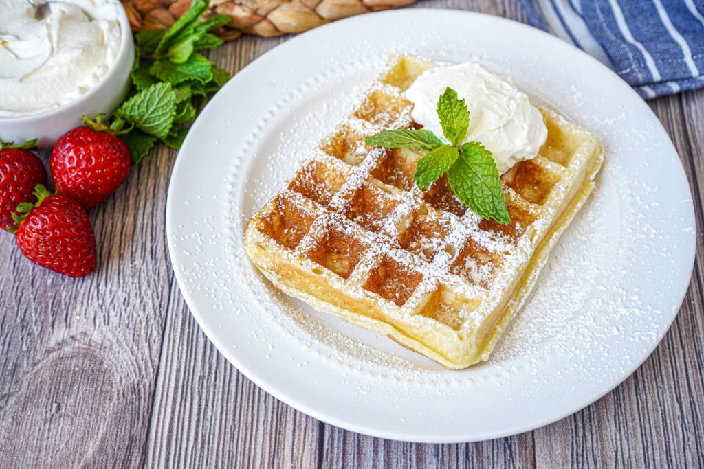 Gaufre de Bruxelles (Brussels Waffle) on a white plate with powdered sugar, fresh mint, and whipped cream.