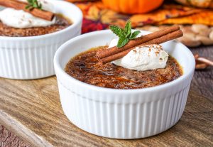Pumpkin Crème Brûlée in two white ramekins with whipped cream and cinnamon stick.