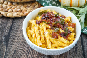Pumpkin and Cider Macaroni and Cheese in a white bowl with bacon and sage.