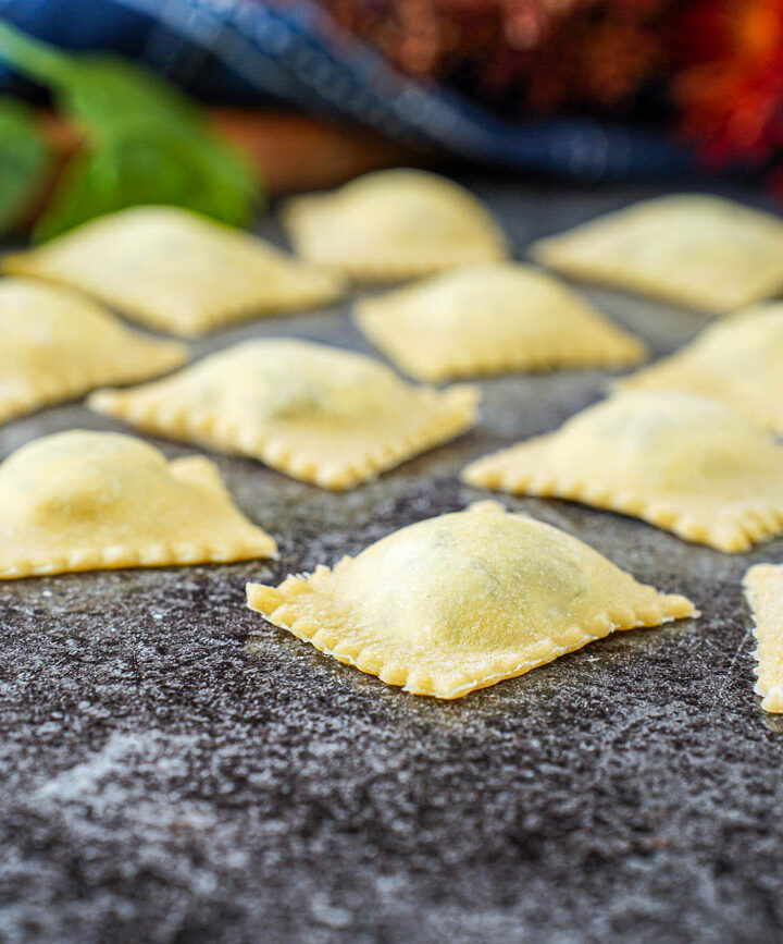 Ravioli Ricotta e Spinaci (Spinach Ricotta Ravioli) on a gray surface with spinach leaves and red flowers in the background.