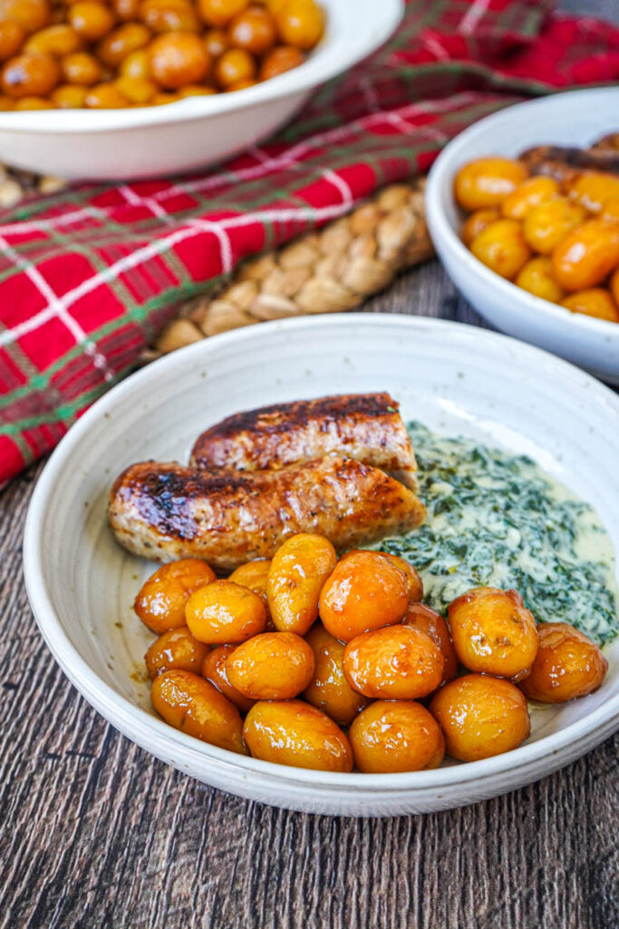 Close up of Brunede Kartofler (Danish Browned Potatoes) on two plates with sausage and creamed kale.