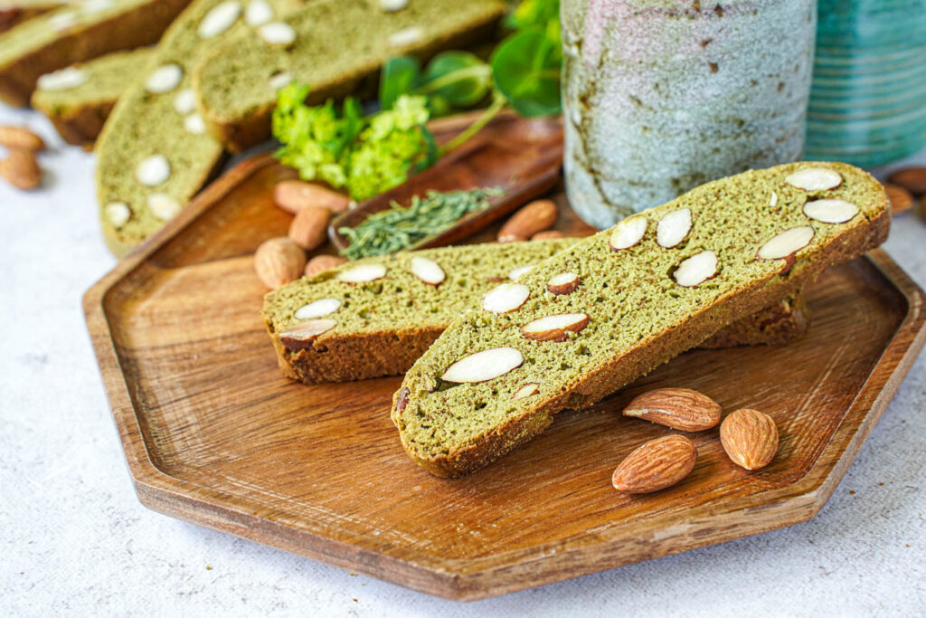 Two Matcha Almond Biscotti on a wooden tray with a tea cup and almonds.
