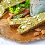 Two Matcha Almond Biscotti on a wooden tray with a tea cup and almonds.