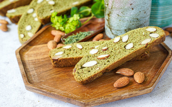 Two Matcha Almond Biscotti on a wooden tray with a tea cup and almonds.