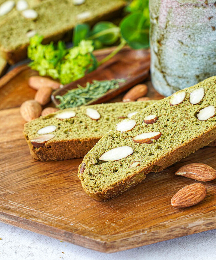 Two Matcha Almond Biscotti on a wooden tray with a tea cup and almonds.