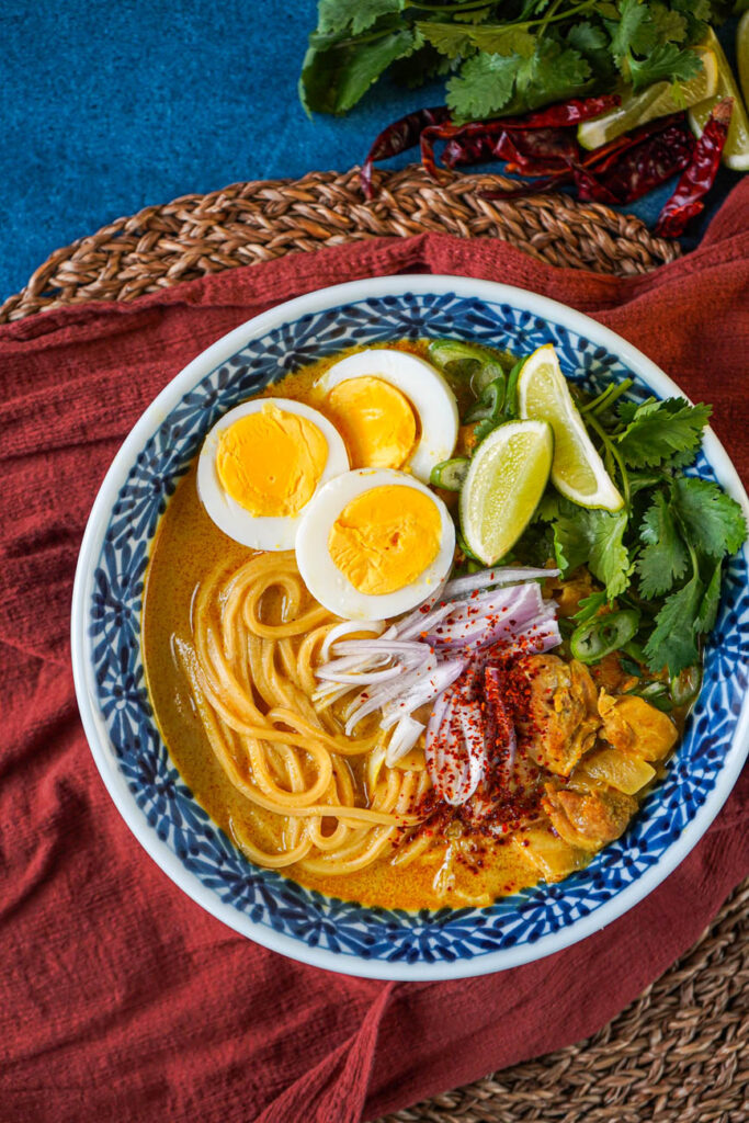 Aerial view of Ohn No Khao Swè (Burmese Chicken Coconut Noodle Soup) in a bowl next to cilantro, lime wedges, and dried chilies.