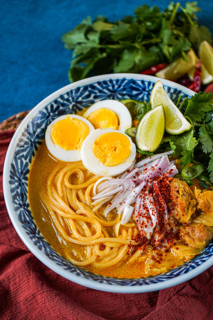 Close up of Ohn No Khao Swè (Burmese Chicken Coconut Noodle Soup) topped with hard boiled eggs, lime wedges, shallots, cilantro, and red chili powder.