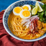 Ohn No Khao Swè (Burmese Chicken Coconut Noodle Soup) in a bowl with hard boiled eggs, lime wedges, cilantro, and thinly sliced shallots.