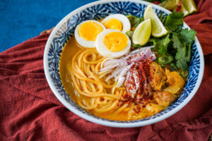 Ohn No Khao Swè (Burmese Chicken Coconut Noodle Soup) in a bowl with hard boiled eggs, lime wedges, cilantro, and thinly sliced shallots.