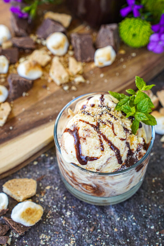 S'mores Ice Cream in a glass bowl next to toasted marshmallows, chopped graham crackers, and chocolate pieces.
