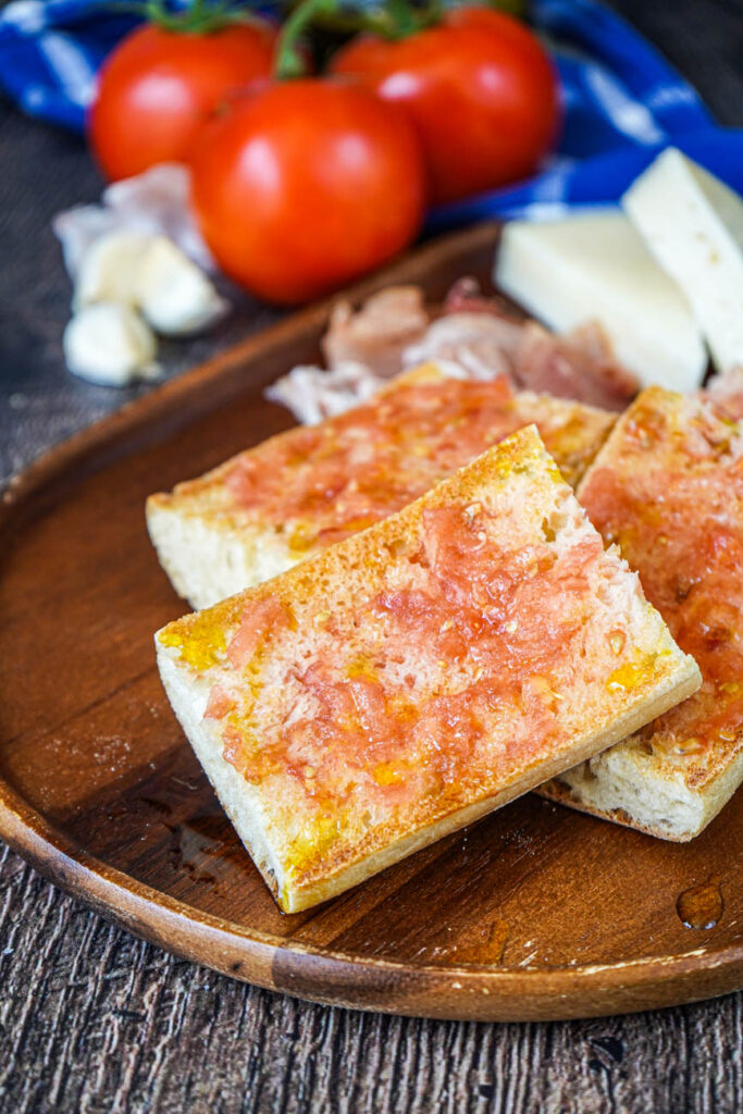 Pa amb Tomàquet (Catalan Bread with Tomato) on a board with tomatoes and garlic in the background.