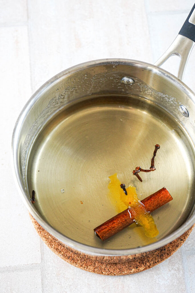Clear syrup in a small saucepan with a cinnamon stick, lemon peel, and cloves.