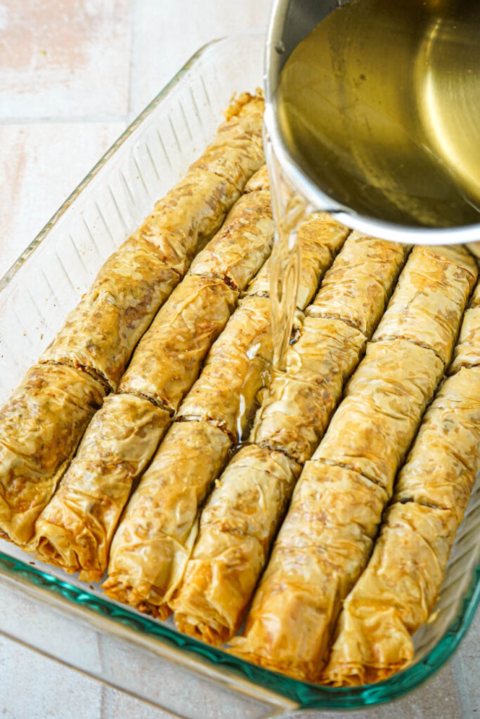 Pouring syrup over Saragli (Greek Rolled Baklava) in a pan.