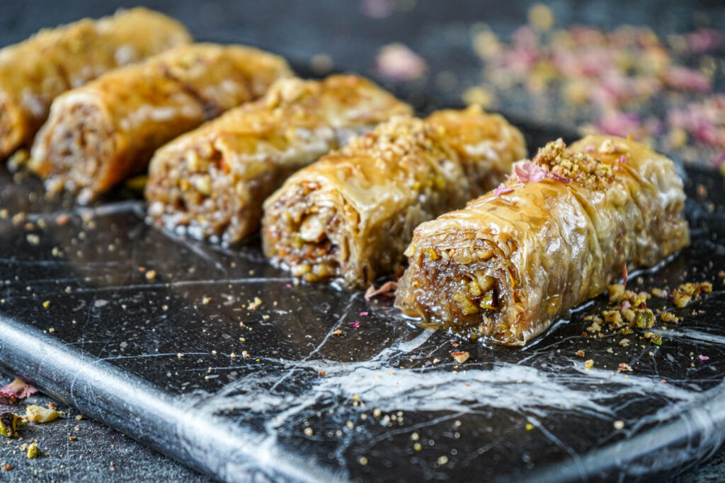 Five Saragli (Greek Rolled Baklava) on a marble board with ground nuts and rose petals over the top.