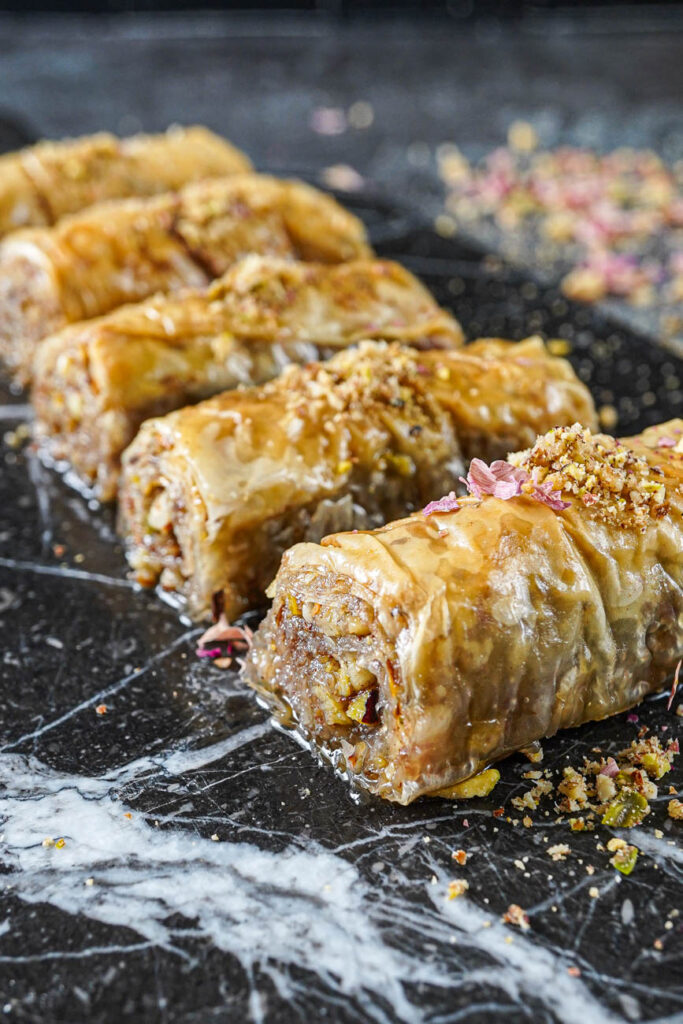Close up of five Saragli (Greek Rolled Baklava) on a marble board with ground nuts and rose petals on top.