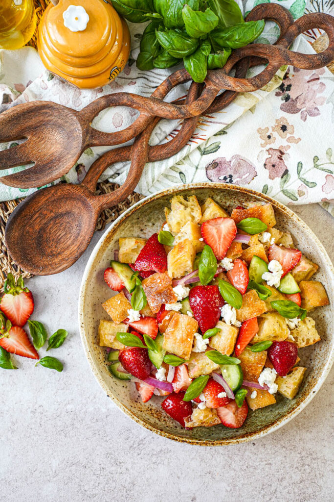 Aerial view of Strawberry Panzanella in a bowl next to strawberry halves, basil leaves, and a wooden spoon.